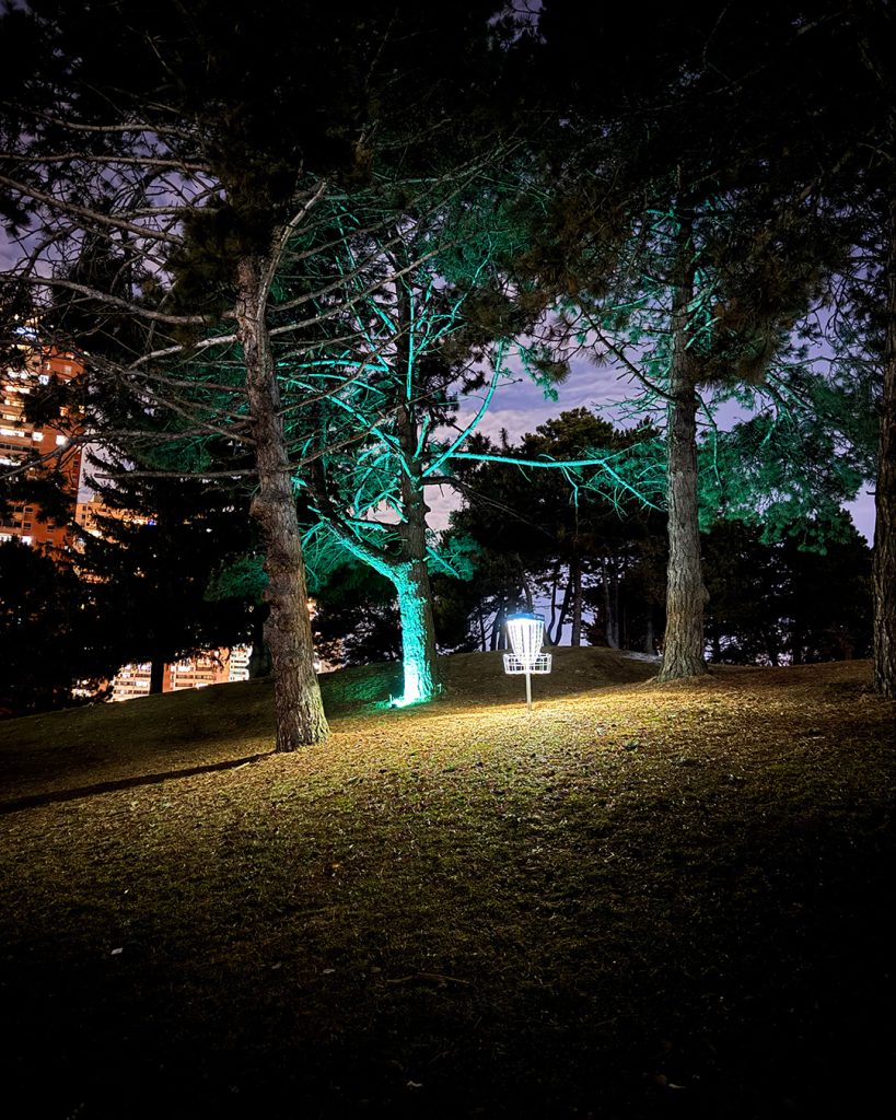 Disc golf basket glowing in the dark surrounded by cyan and green light during a Dr. Glow nighttime disc golf experience in Toronto