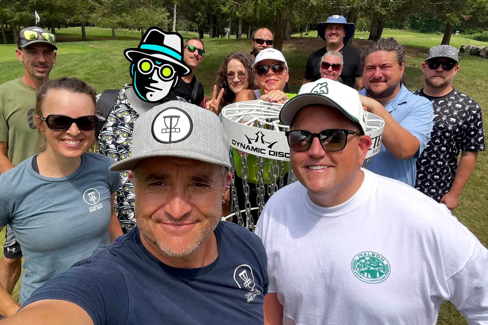 Cara and Jeff from ChainLink Disc Golf and Cale from Black Bear Ridge take a group selfie with Dr. Glow and players gathered around the disc golf basket in daylight.