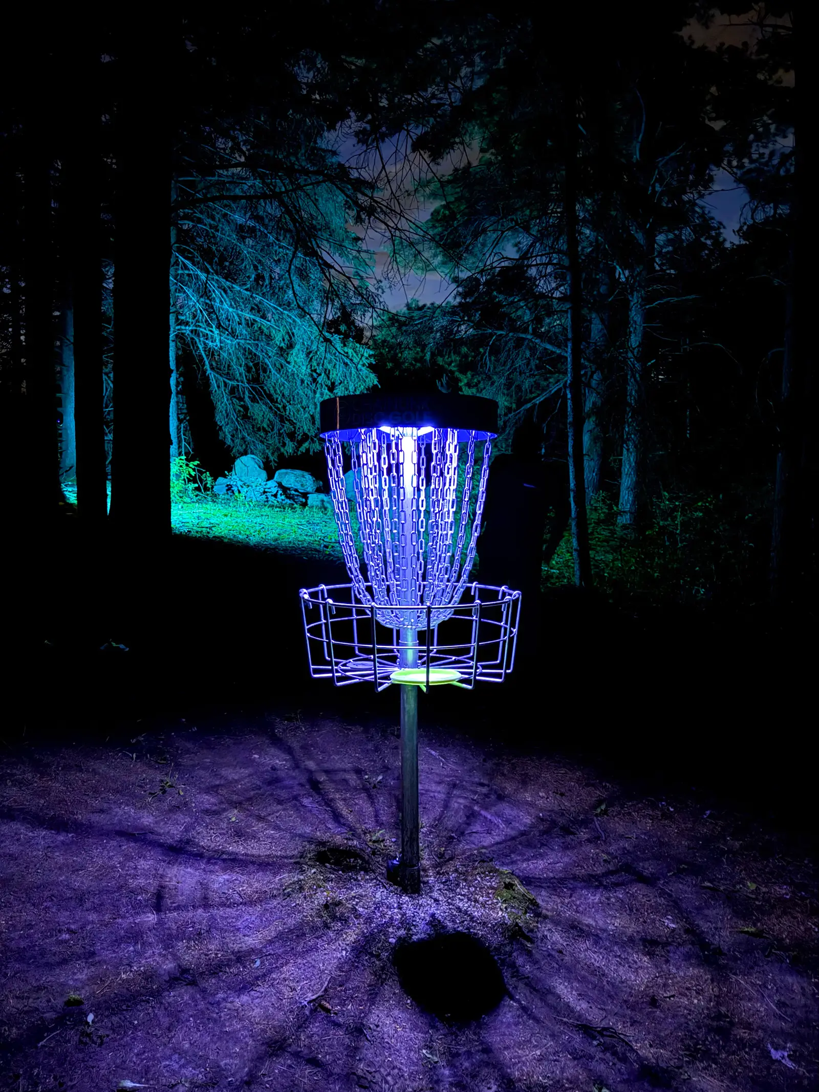 A disc golf basket illuminated by blue UV light surrounded by dark forest trees at the Glow for More event at Albion Hills