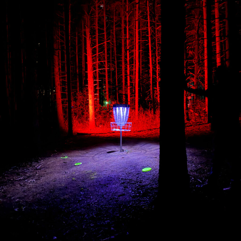 A disc golf basket surrounded by red lit trees with three glowing discs on the ground at the Glow for More event at Albion Hills Conservation Area