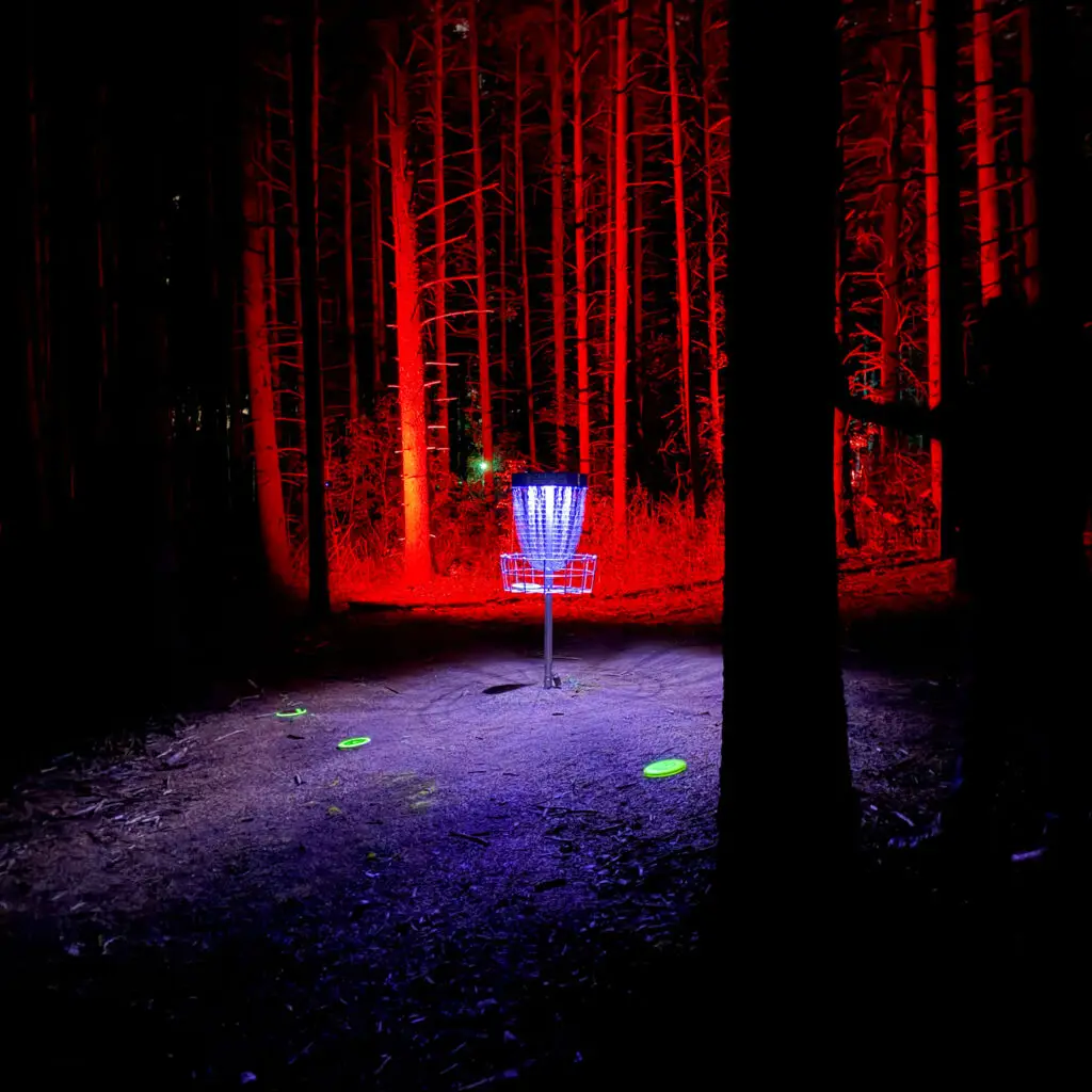 A disc golf basket surrounded by red lit trees with three glowing discs on the ground at the Glow for More event at Albion Hills Conservation Area