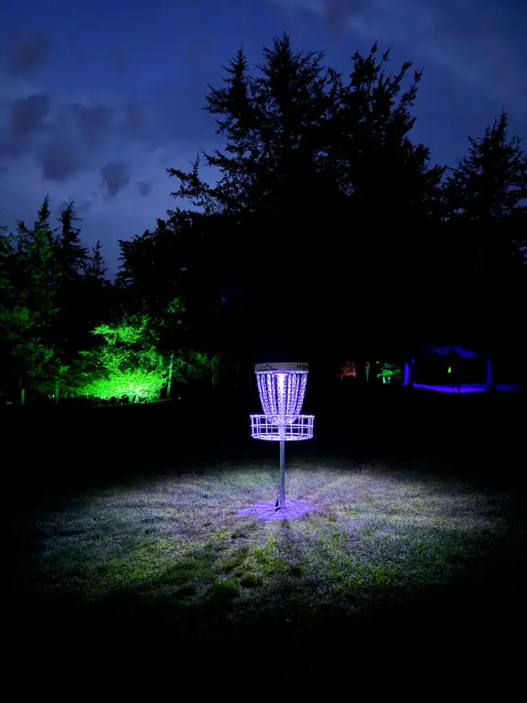 A disc golf basket glows purple in an open grassy field at night, framed by green and blue lit trees at Black Bear Ridge.