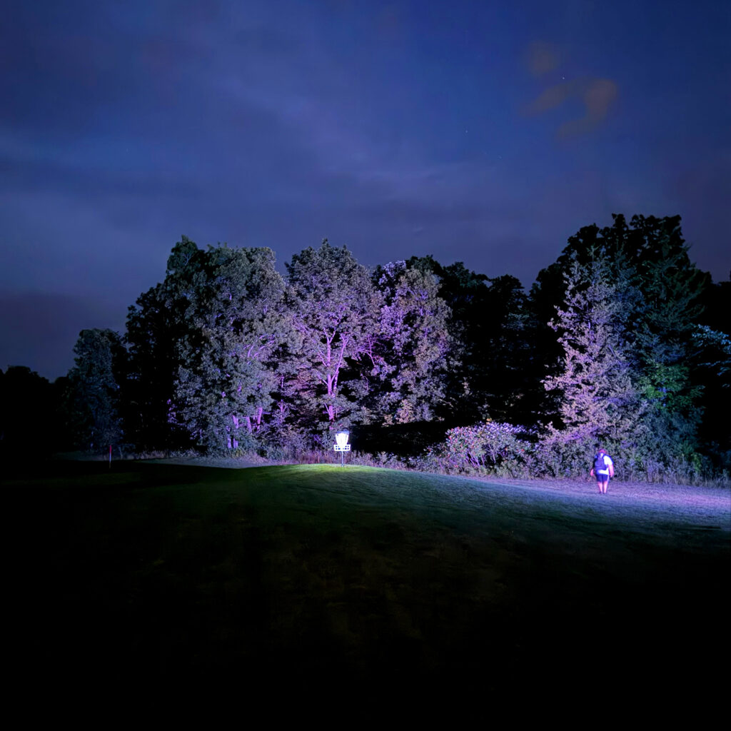 A player walks toward a glowing disc golf basket at dusk on the Forest and Fairways course at Black Bear Ridge, trees lit purple in the background.