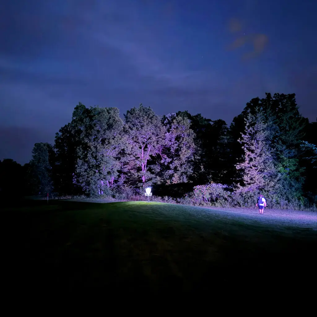 A player walks toward a glowing disc golf basket at dusk on the Forest and Fairways course at Black Bear Ridge, trees lit purple in the background.