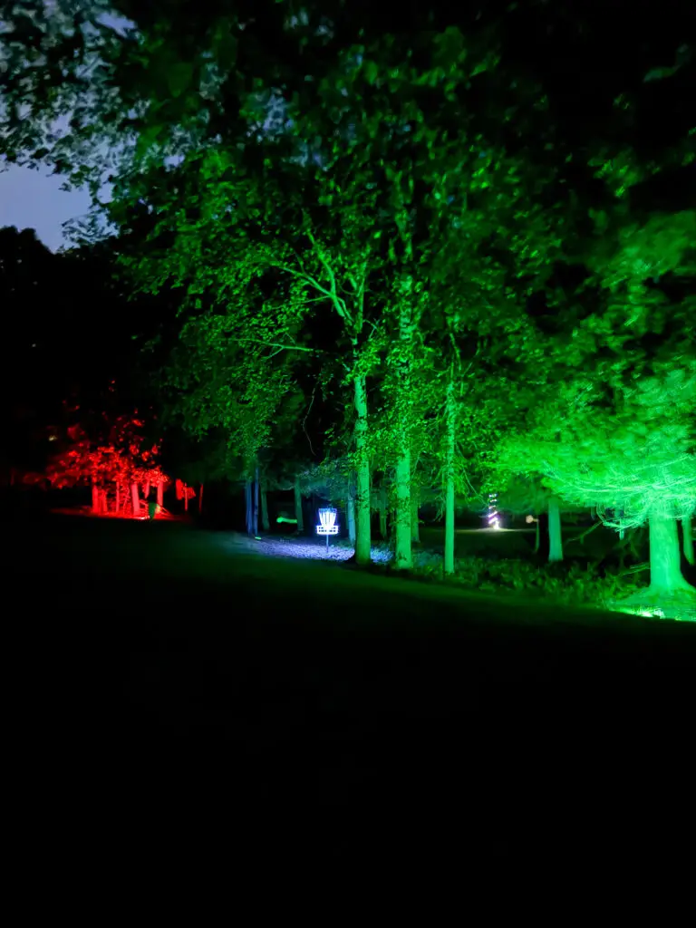 A disc golf basket glows blue in the distance through a forest corridor lit in red and green at Black Bear Ridge.