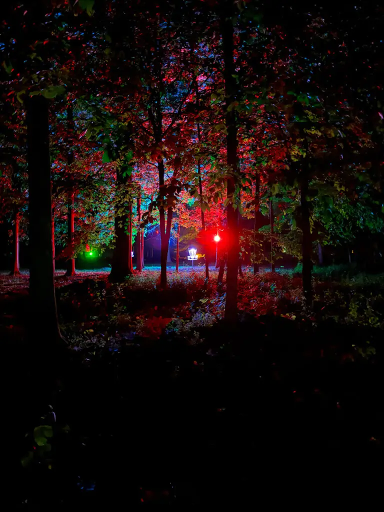A disc golf basket glows white deep in a forest lit with vivid red and green uplighting at the Forest and Fairways glow round.