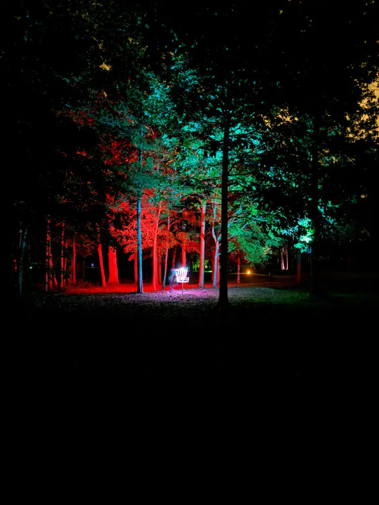 A glowing disc golf basket stands among tree trunks lit red on one side and teal on the other at the Black Bear Ridge glow round.