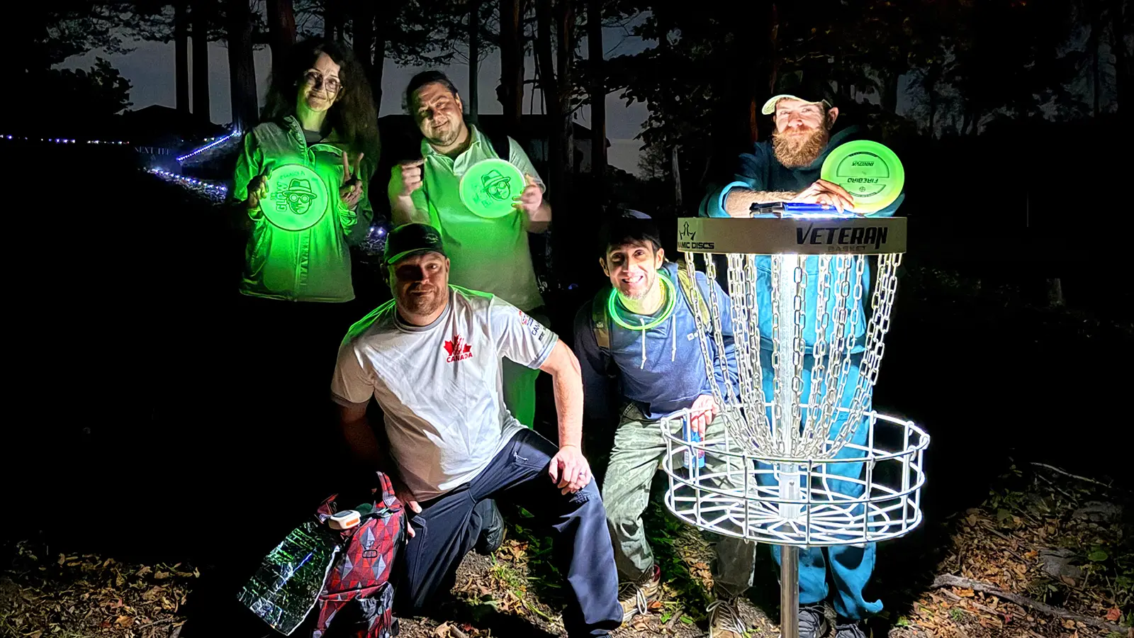 Five players holding glowing green Dr. Glow branded discs beside a lit-up disc golf basket at night.