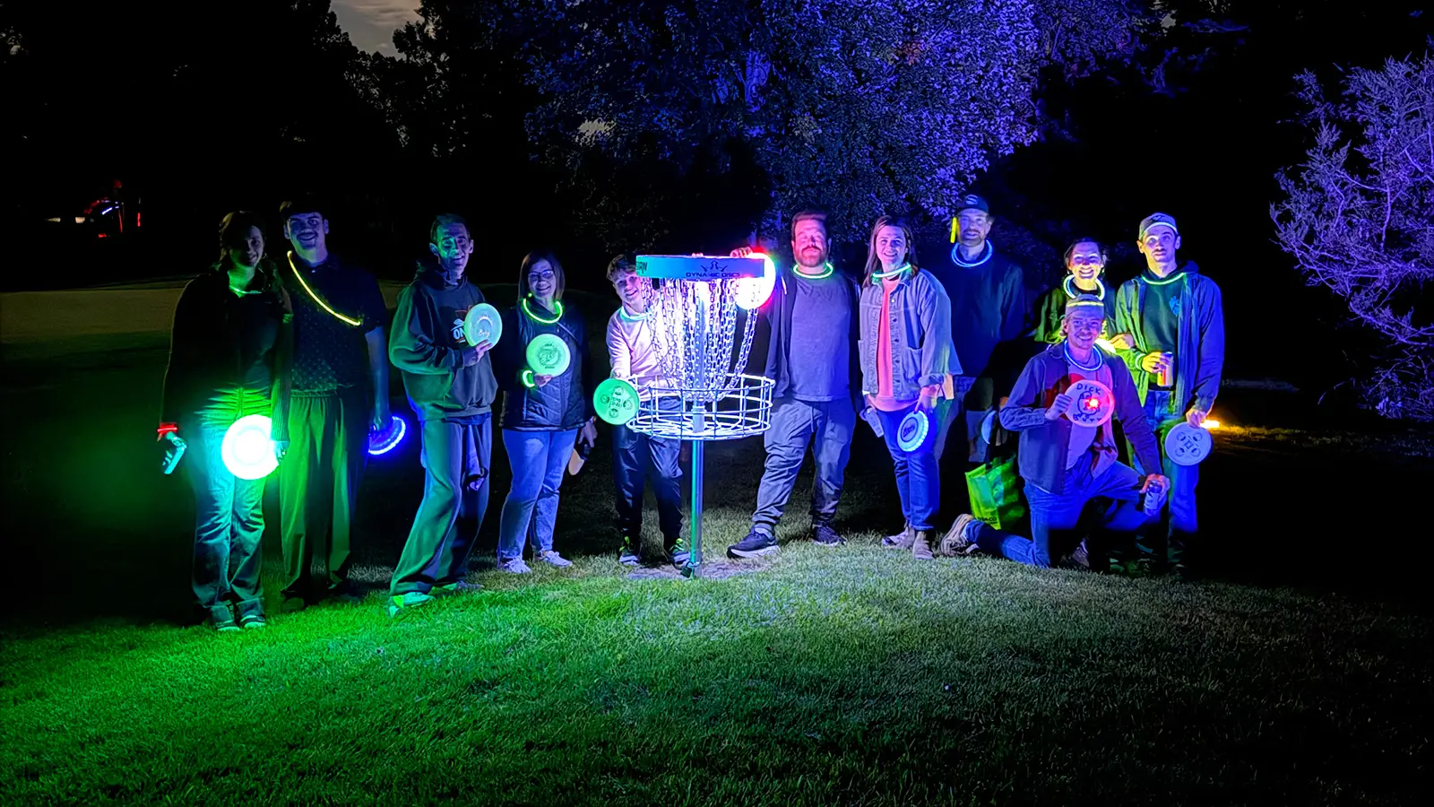 Large group of glow disc golfers, including a family playing for the first time, gathered around an illuminated basket at night.