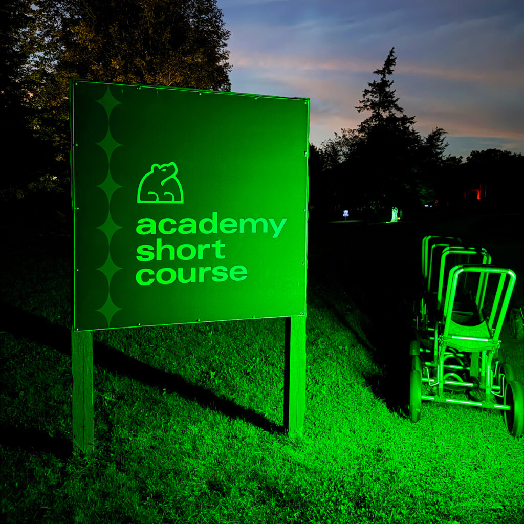 Illuminated green "Academy Short Course" sign with the Black Bear Ridge bear logo, disc carts beside it at dusk.