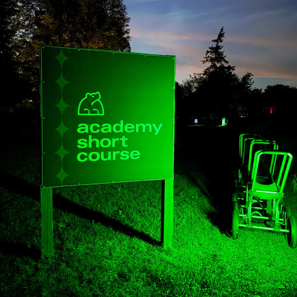 Illuminated green "Academy Short Course" sign with the Black Bear Ridge bear logo, disc carts beside it at dusk.