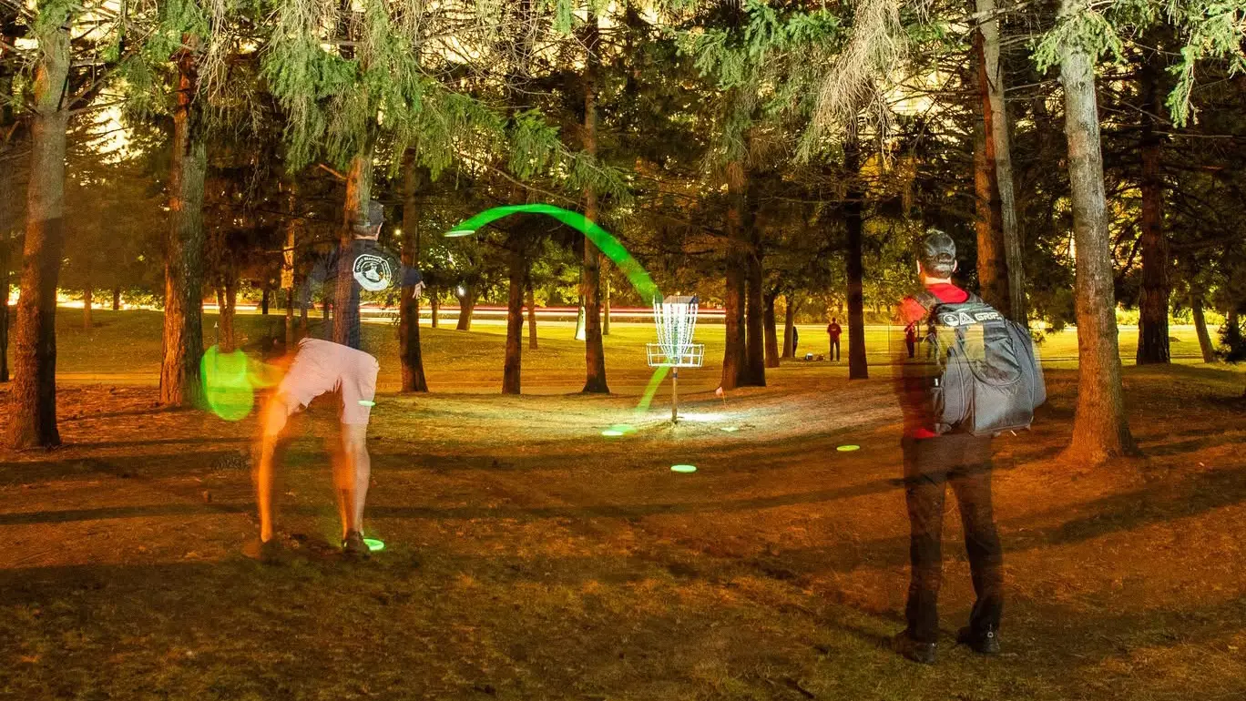Glow disc golf disc in flight toward a lit basket surrounded by trees at night