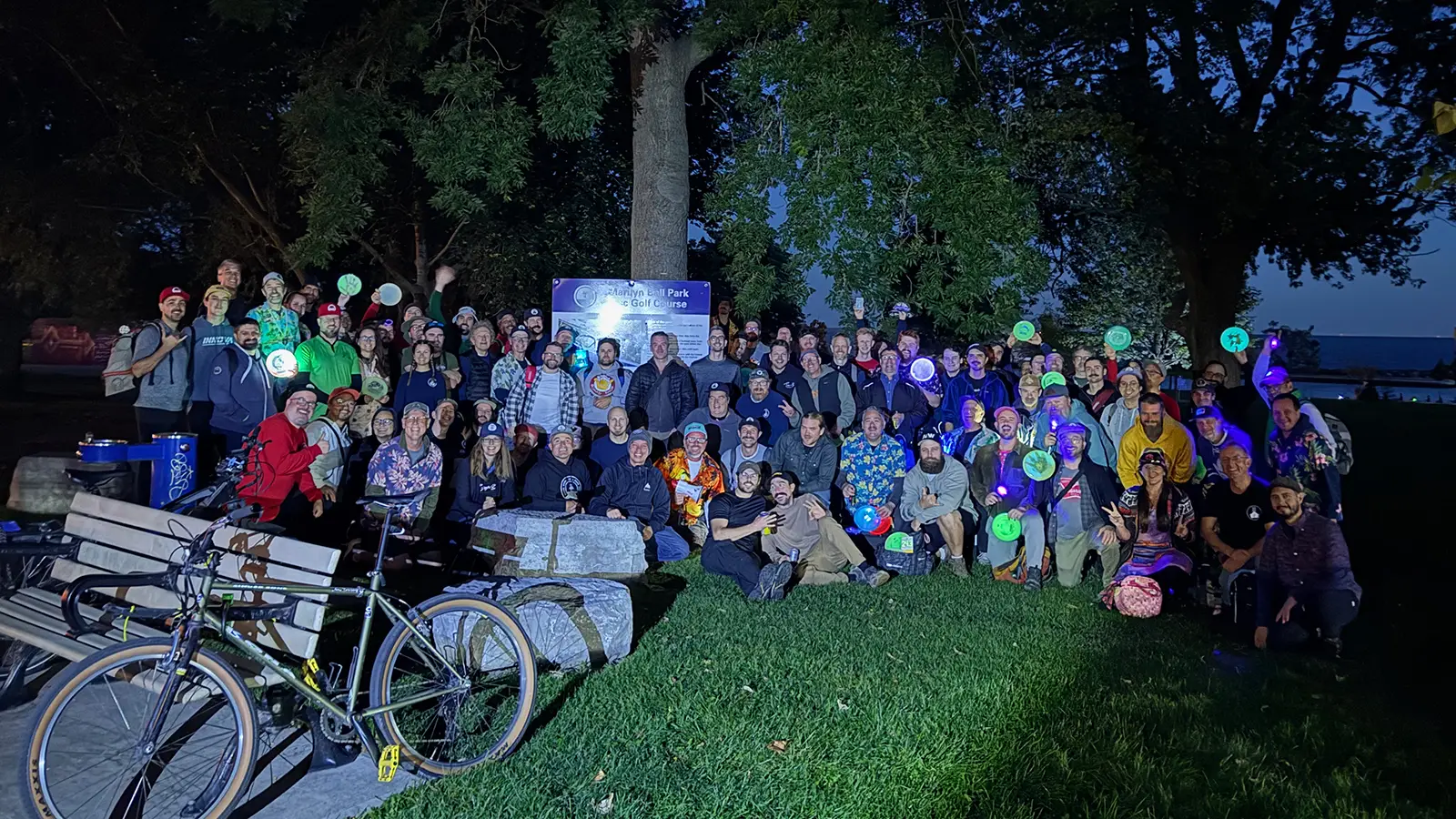 Large group of disc golfers holding glow discs at Marilyn Bell Park Disc Golf Course at dusk