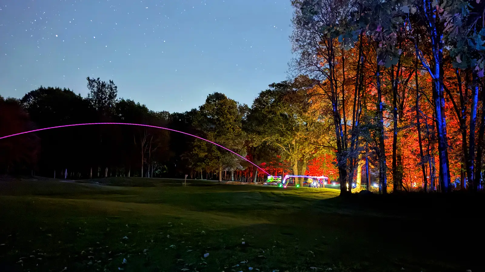 A pink LED golf ball trail arcs across a starry night sky above Black Bear Ridge's Academy Short Course, with red and blue lit trees in the distance.