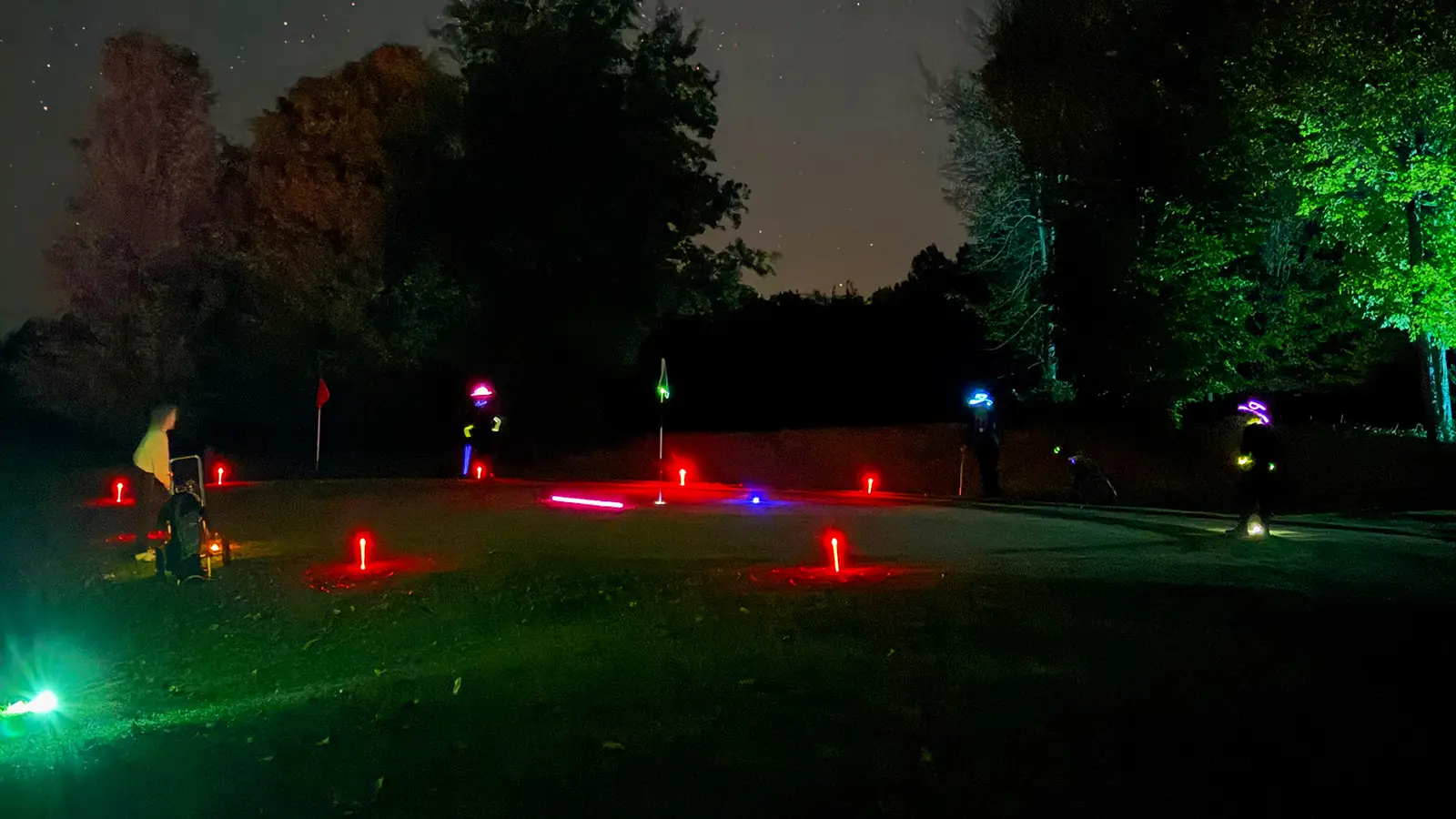 A group of players wearing glow necklaces and hats stand around a green ringed with red LED tee markers under a starry sky at Black Bear Ridge.