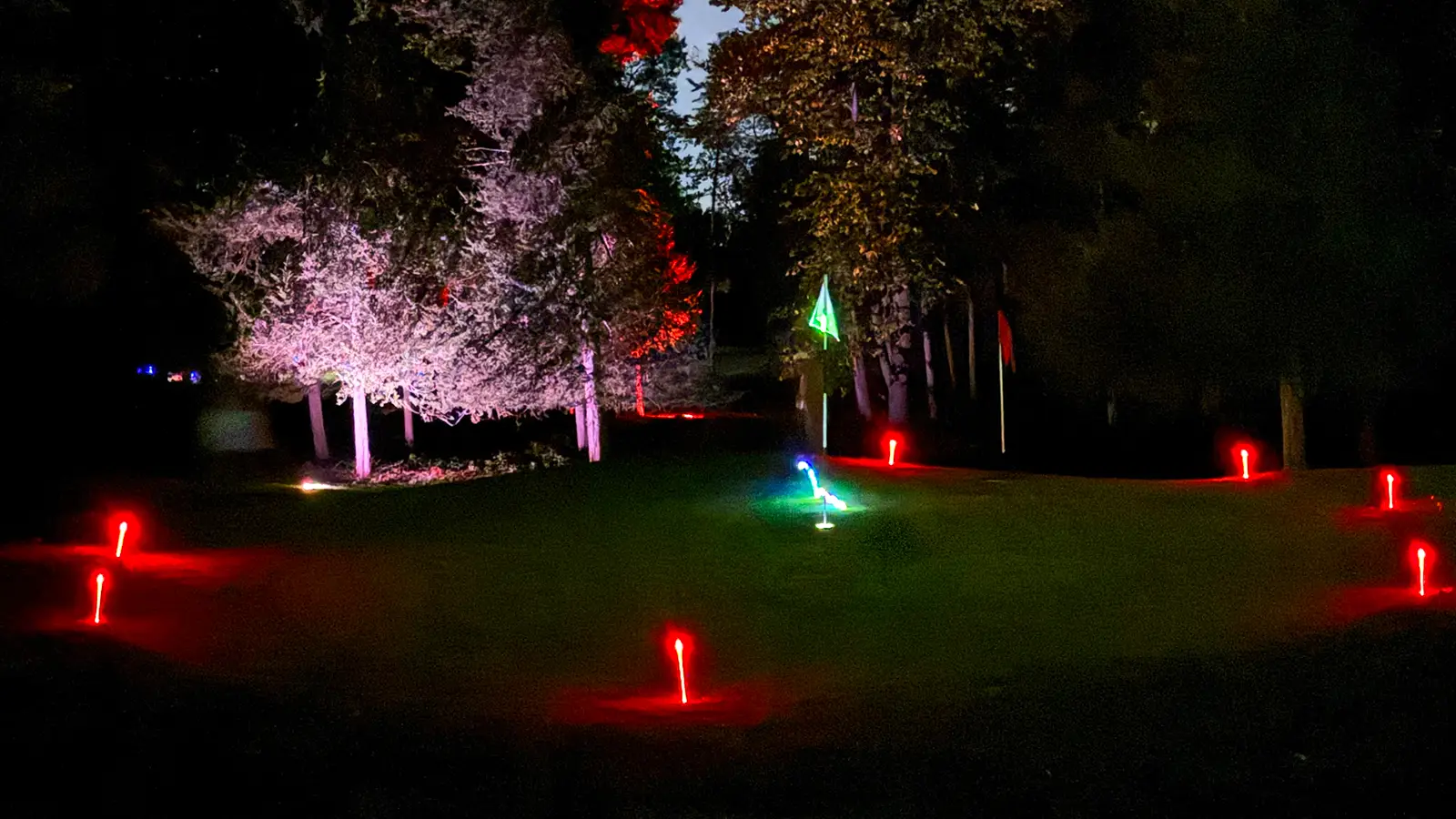 A green glow flag marks the hole on a Black Bear Ridge green surrounded by red LED tee markers, with a white-lit frosted tree and a red-lit tree behind it.