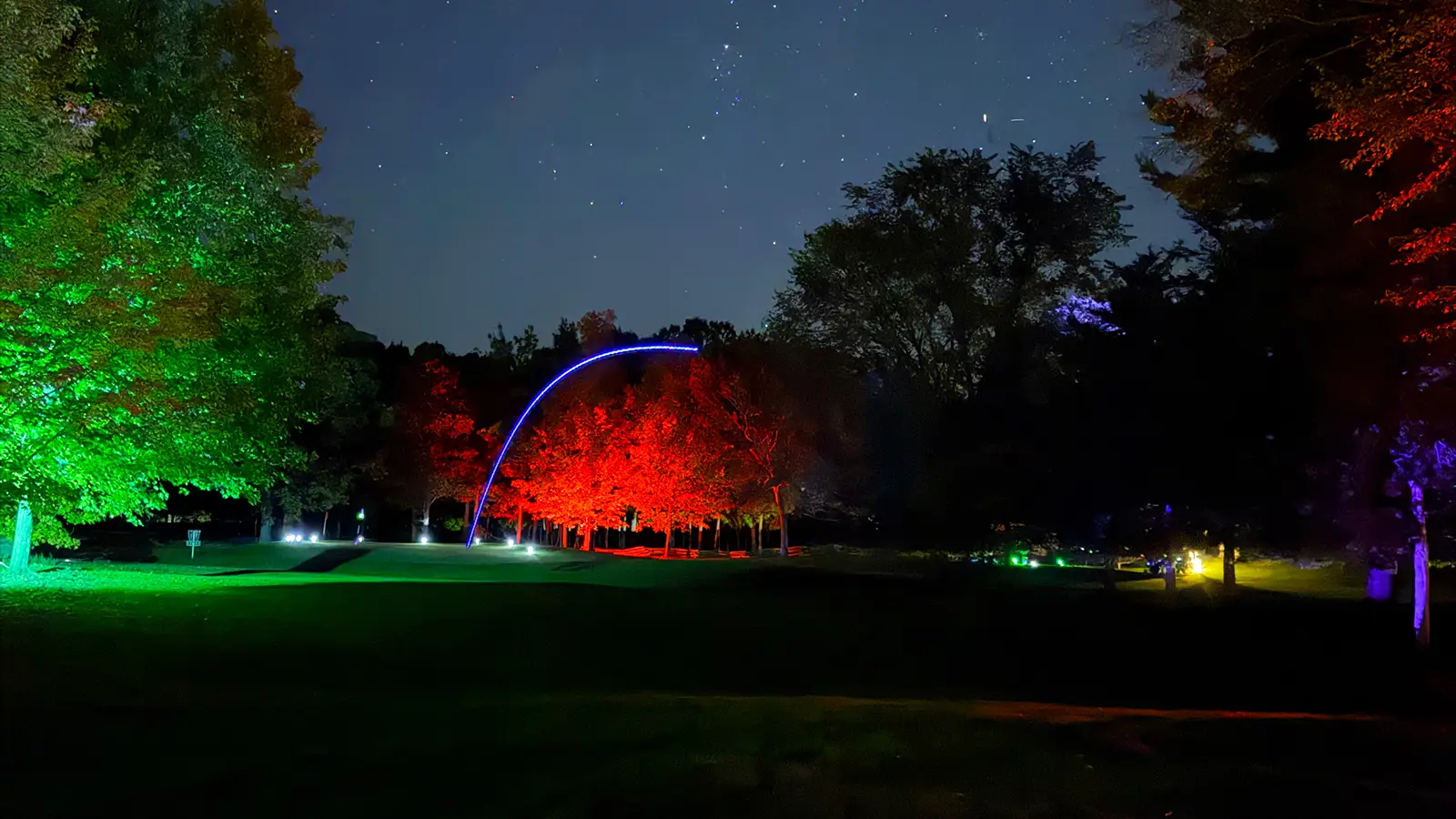 A blue LED golf ball trail arcs through a starry sky between trees lit green on one side and red on the other at Black Bear Ridge.