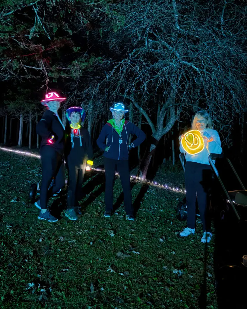 A group including a child wears glowing LED hats in pink, blue, and teal while standing near string lights wrapped around a fallen log in a wooded area.
