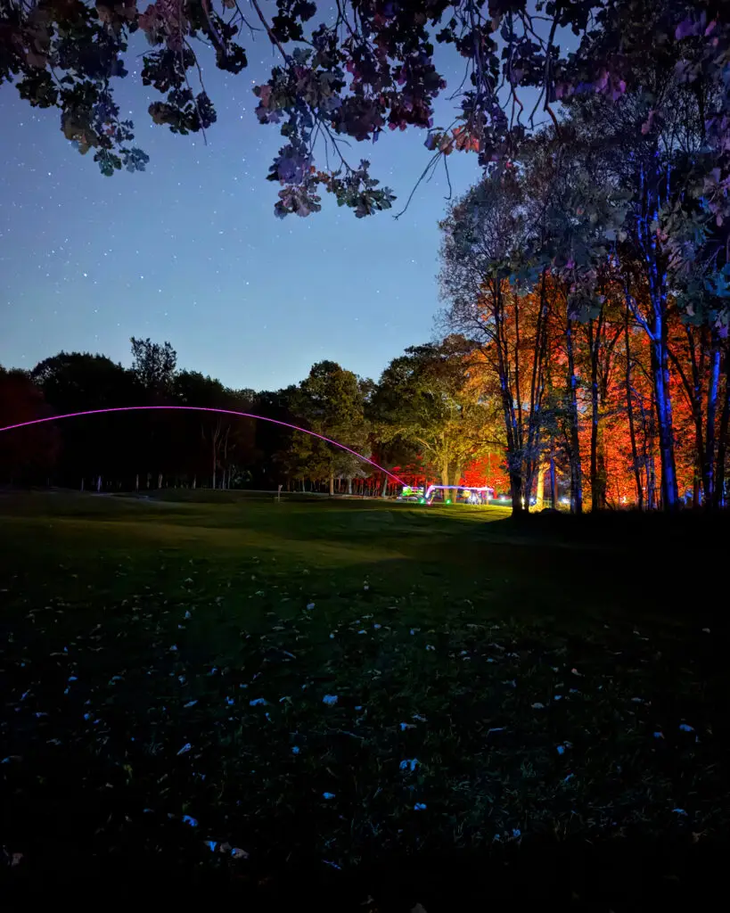 A pink LED ball trail arcs over a brightly lit gathering area at Black Bear Ridge, with orange and red lit trees and a starry sky above.