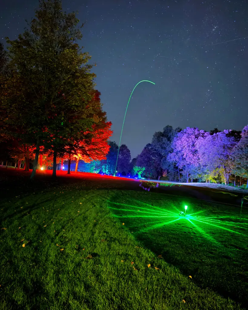 A green LED ball trail arcs high into a starry night sky between a red-lit tree on the left and purple-lit trees on the right.