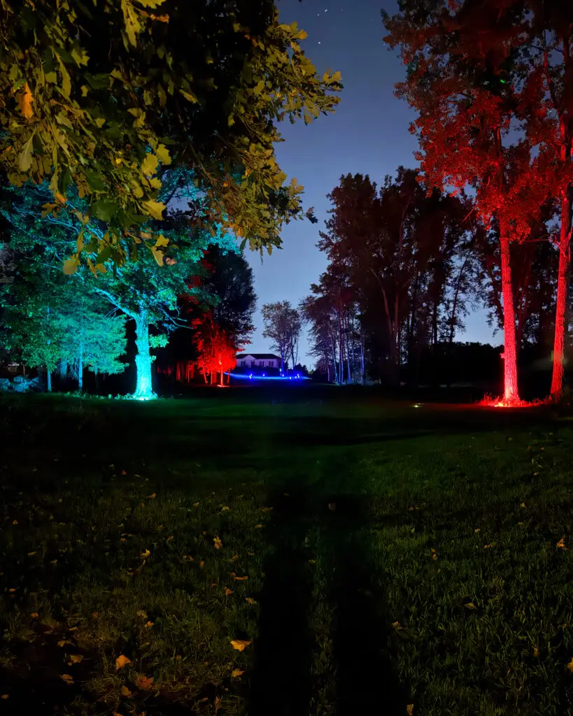 A teal-lit tree and red-lit trees frame a fairway lined with blue LED markers, with a house visible in the distance under a starry sky.