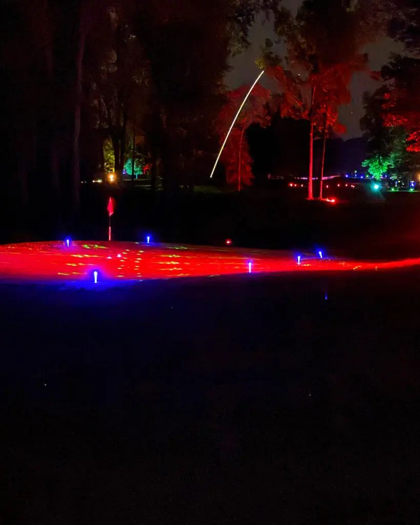 A golf ball's glowing trail arcs over a red-lit tree line toward a green marked with a red flag and blue LED tee markers.