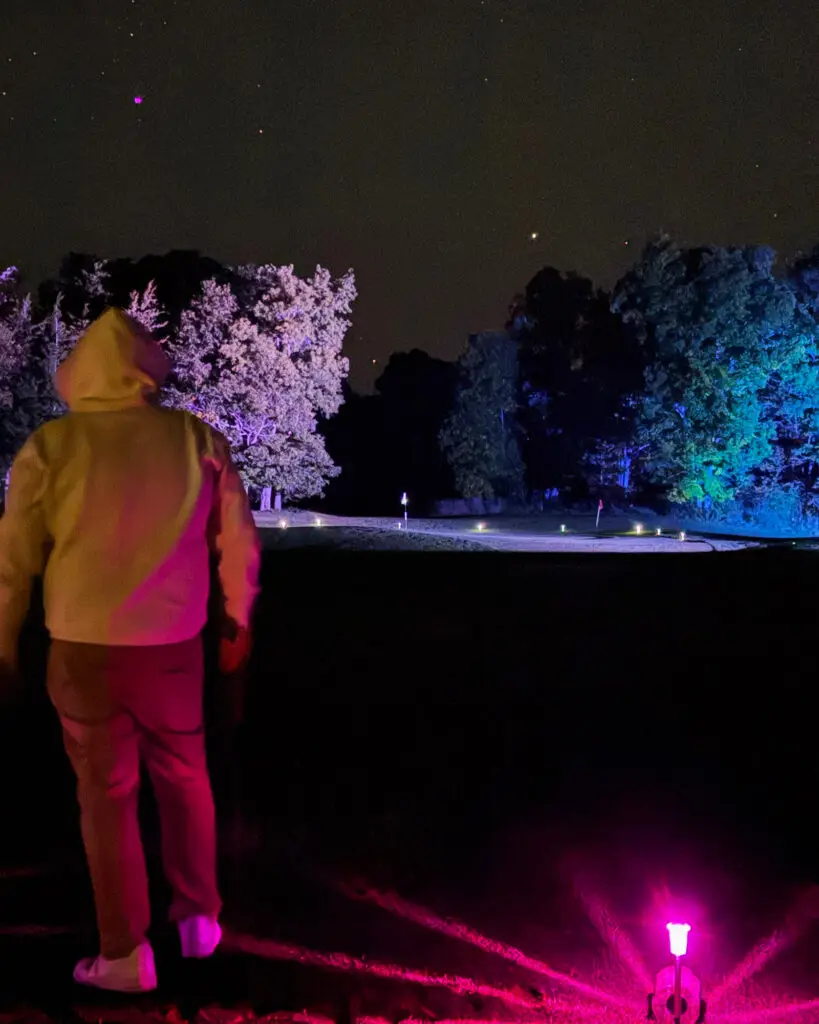 A player in a glowing pink and yellow hoodie looks out over a lit fairway at Black Bear Ridge, with frosted white and blue lit trees in the distance.