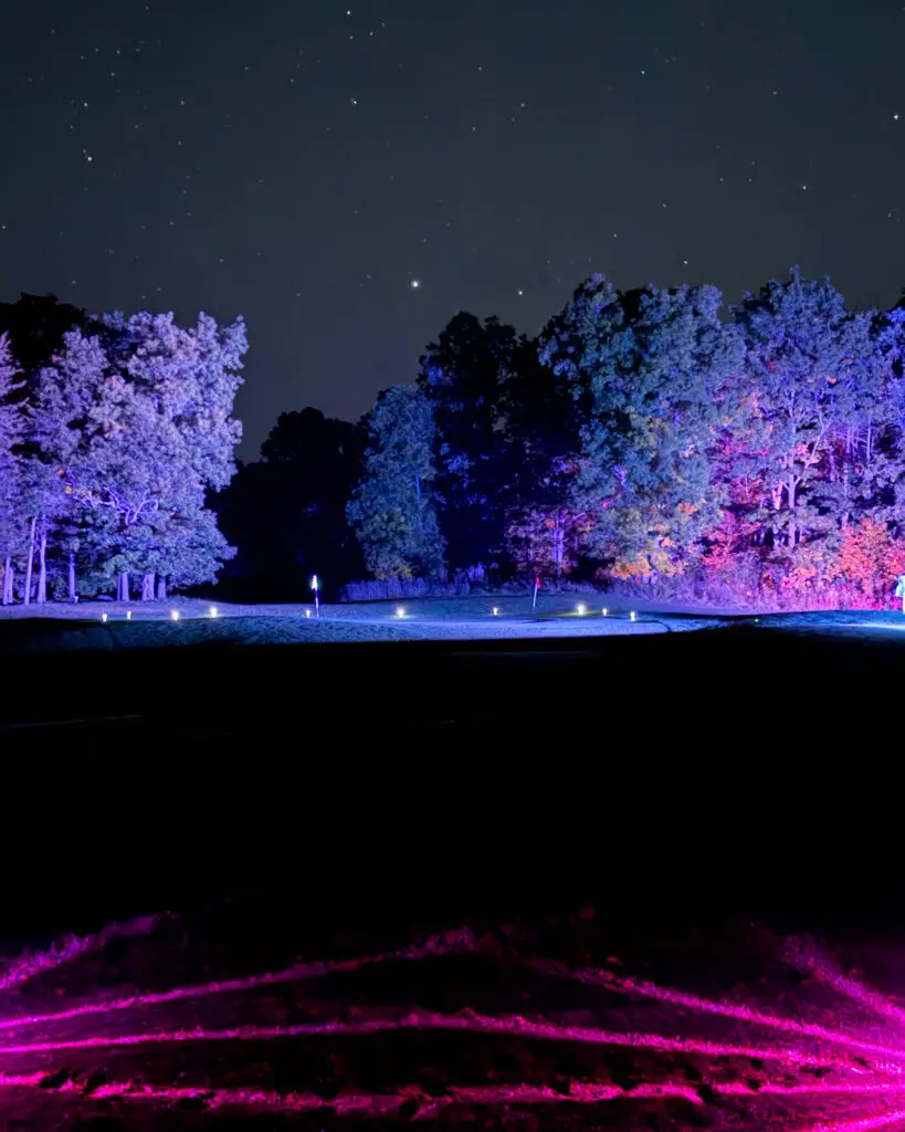 A Black Bear Ridge green glows under purple-lit trees and blue LED tee markers, with a red flag marking the hole and pink light streaking across the foreground grass.