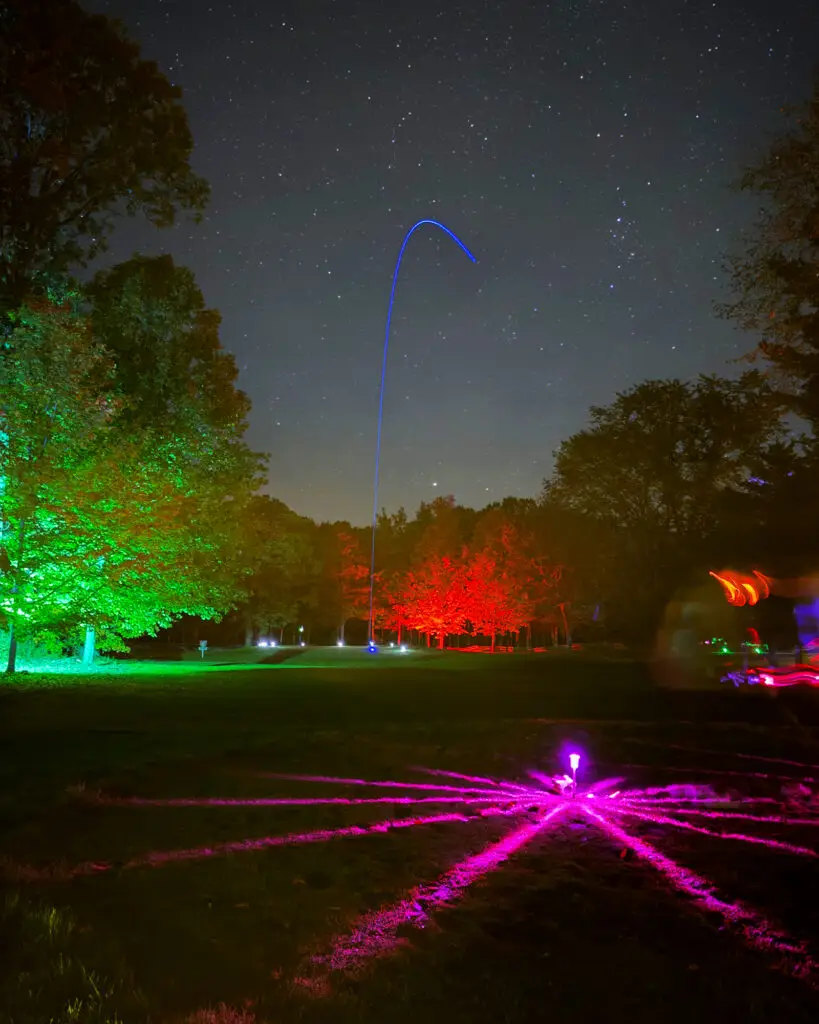 A blue LED golf ball trail arcs high into a starry sky above trees lit green and red at Black Bear Ridge's Academy Short Course.