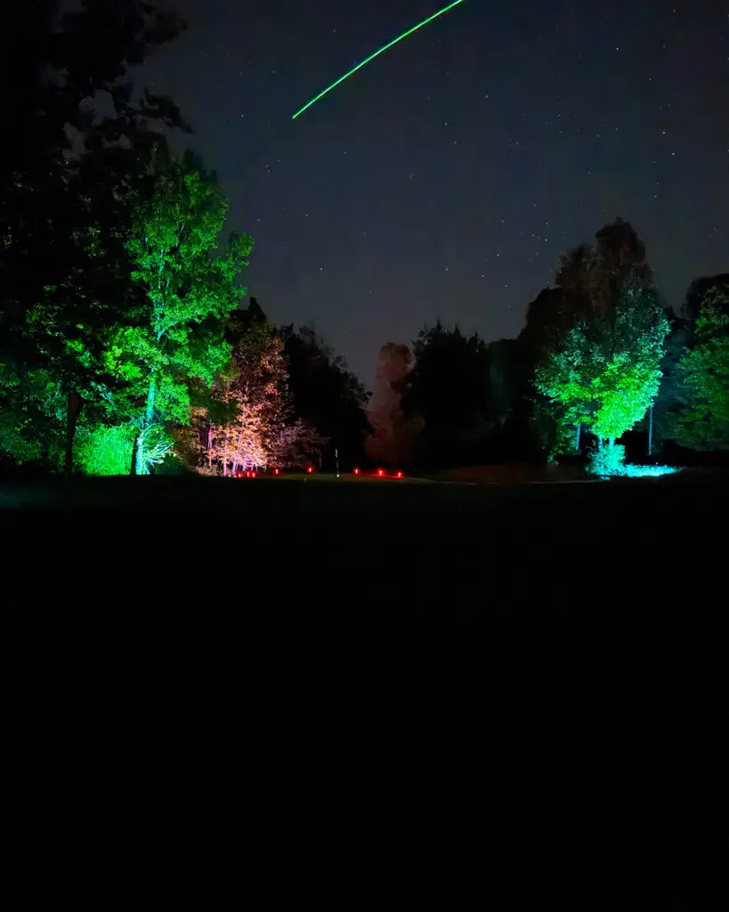 A green LED golf ball trail streaks across a starry sky above a green ringed with red LED tee markers and trees lit in green, pink, and blue.