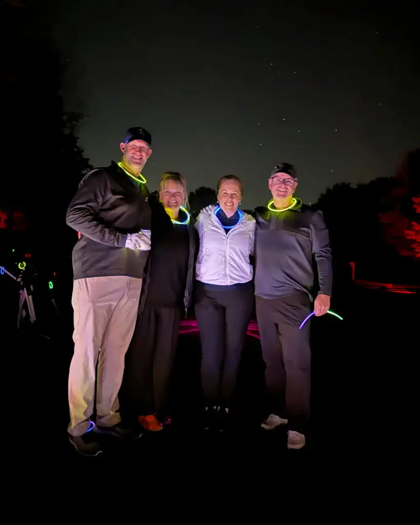 Four smiling golfers wearing glow necklaces pose together at night, with pull carts and red-lit trees behind them under a starry sky.