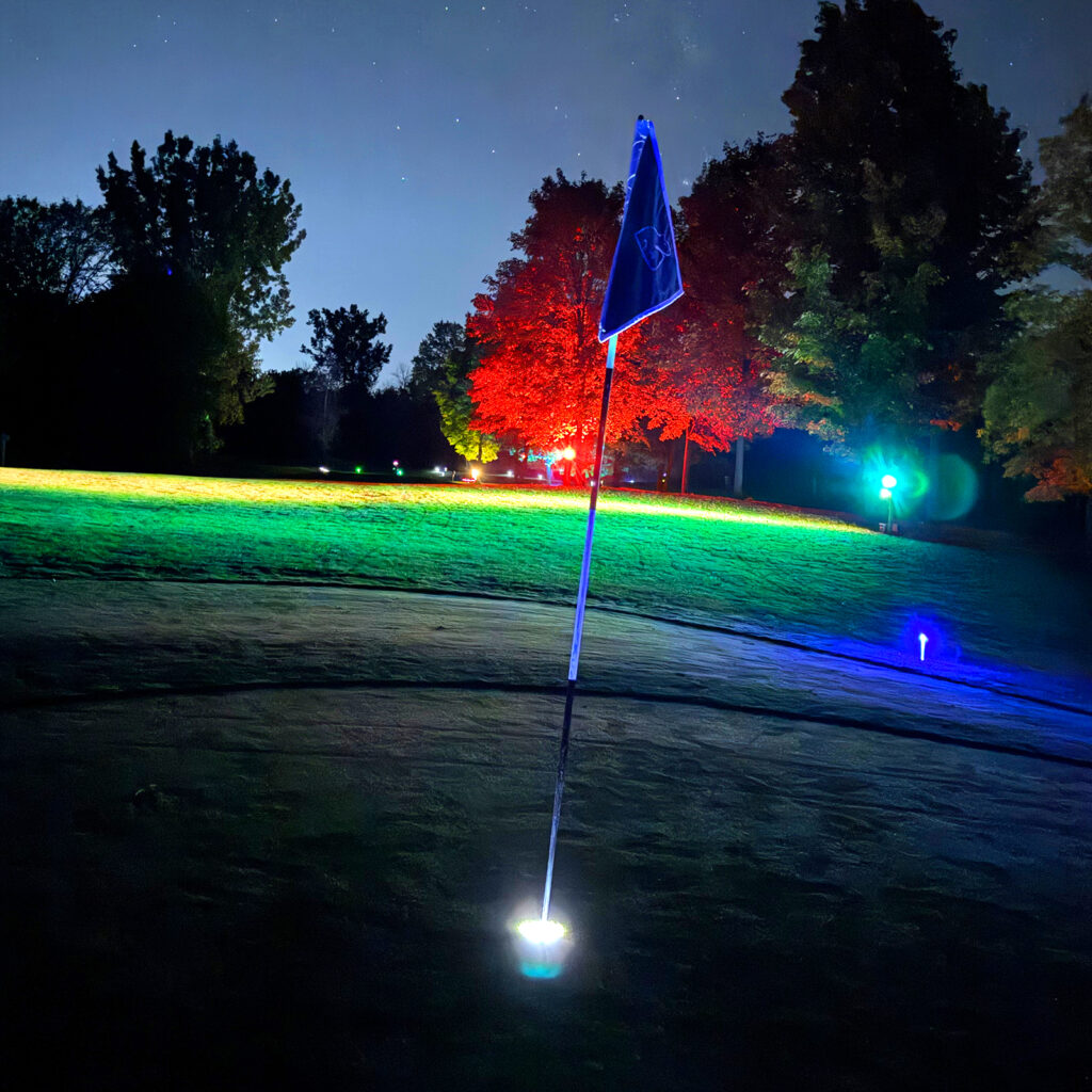 A glowing blue flagstick on a Black Bear Ridge green at night, lit by a white base light, with red and green lit trees and a starry sky behind it.
