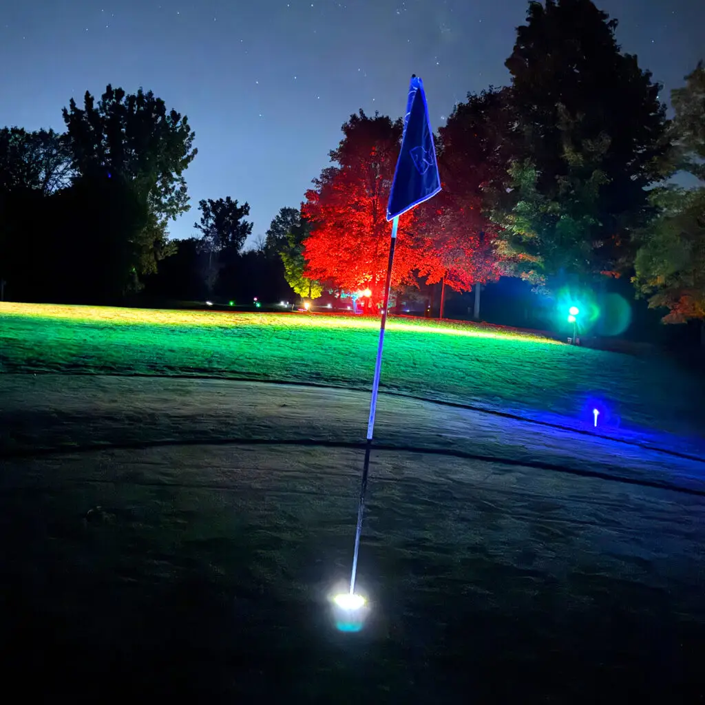 A glowing blue flagstick on a Black Bear Ridge green at night, lit by a white base light, with red and green lit trees and a starry sky behind it.