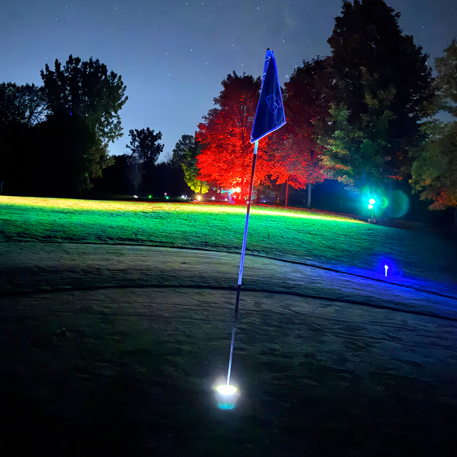 A glowing blue flagstick on a Black Bear Ridge green at night, lit by a white base light, with red and green lit trees and a starry sky behind it.