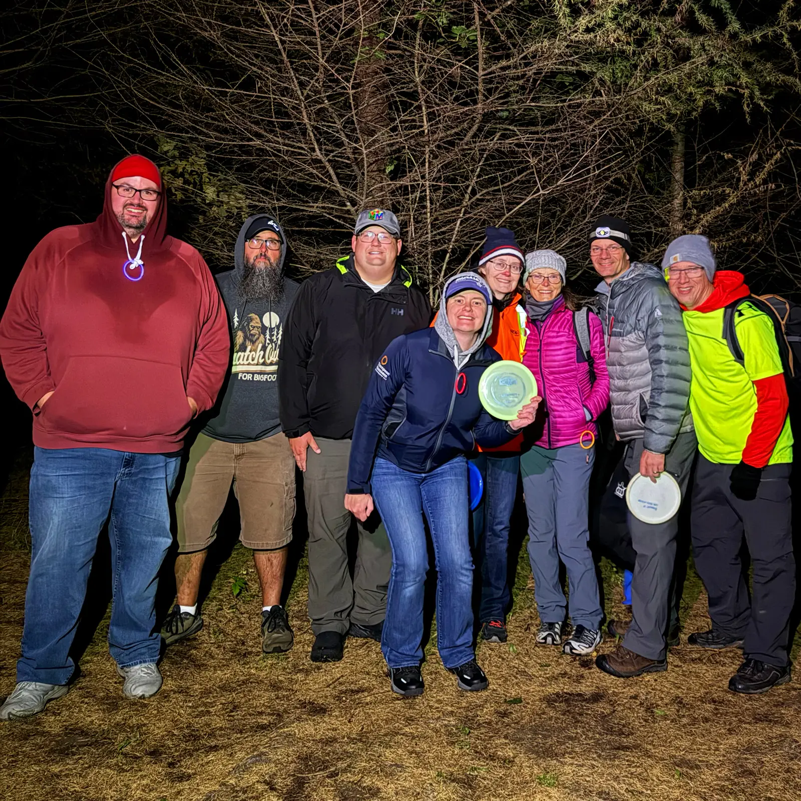 Group of GLOWLOT players smiling together in the dark woods at GoodLot, several holding glow discs