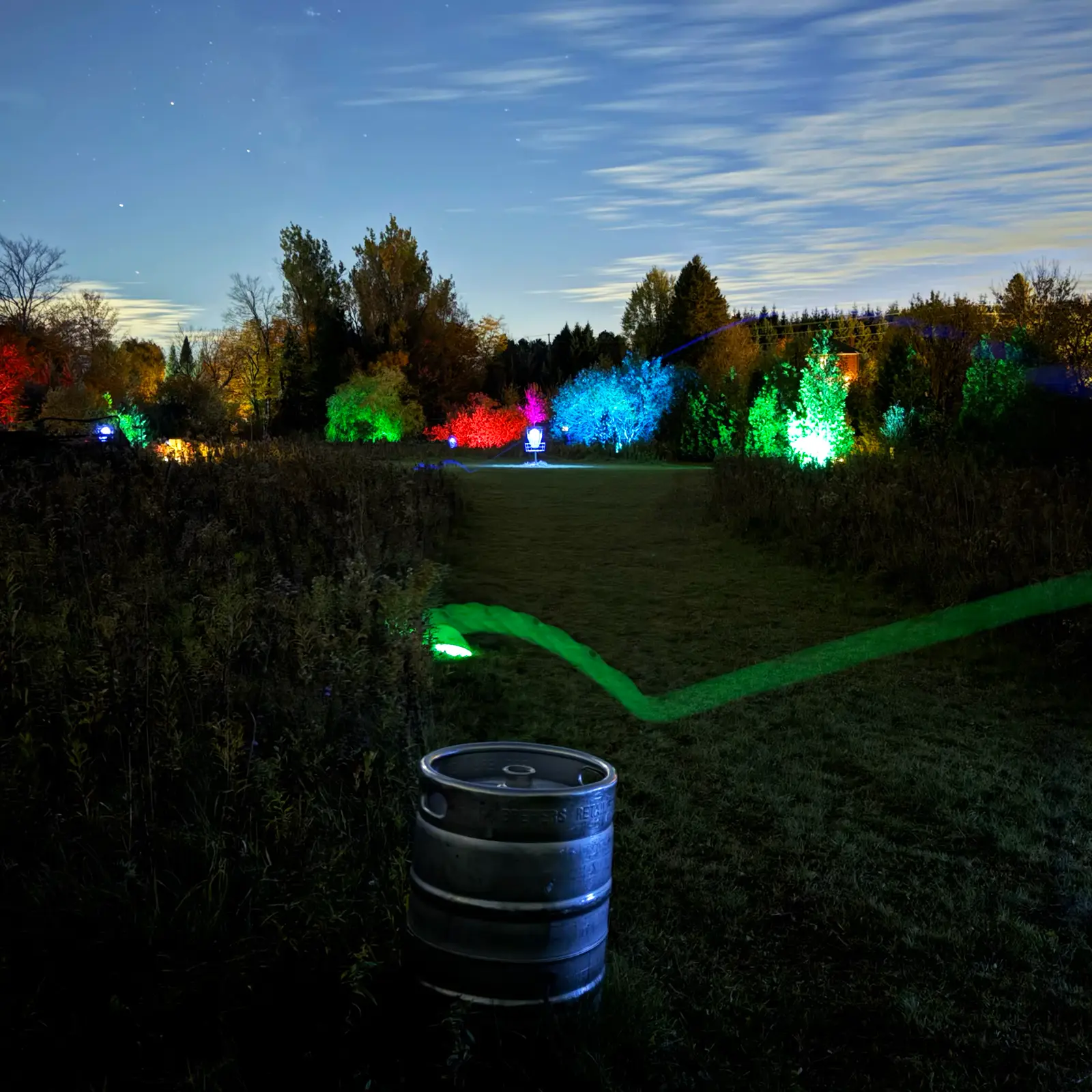 A beer keg glowing in the foreground with a rainbow of lit-up trees and a disc golf basket in the distance at GLOWLOT