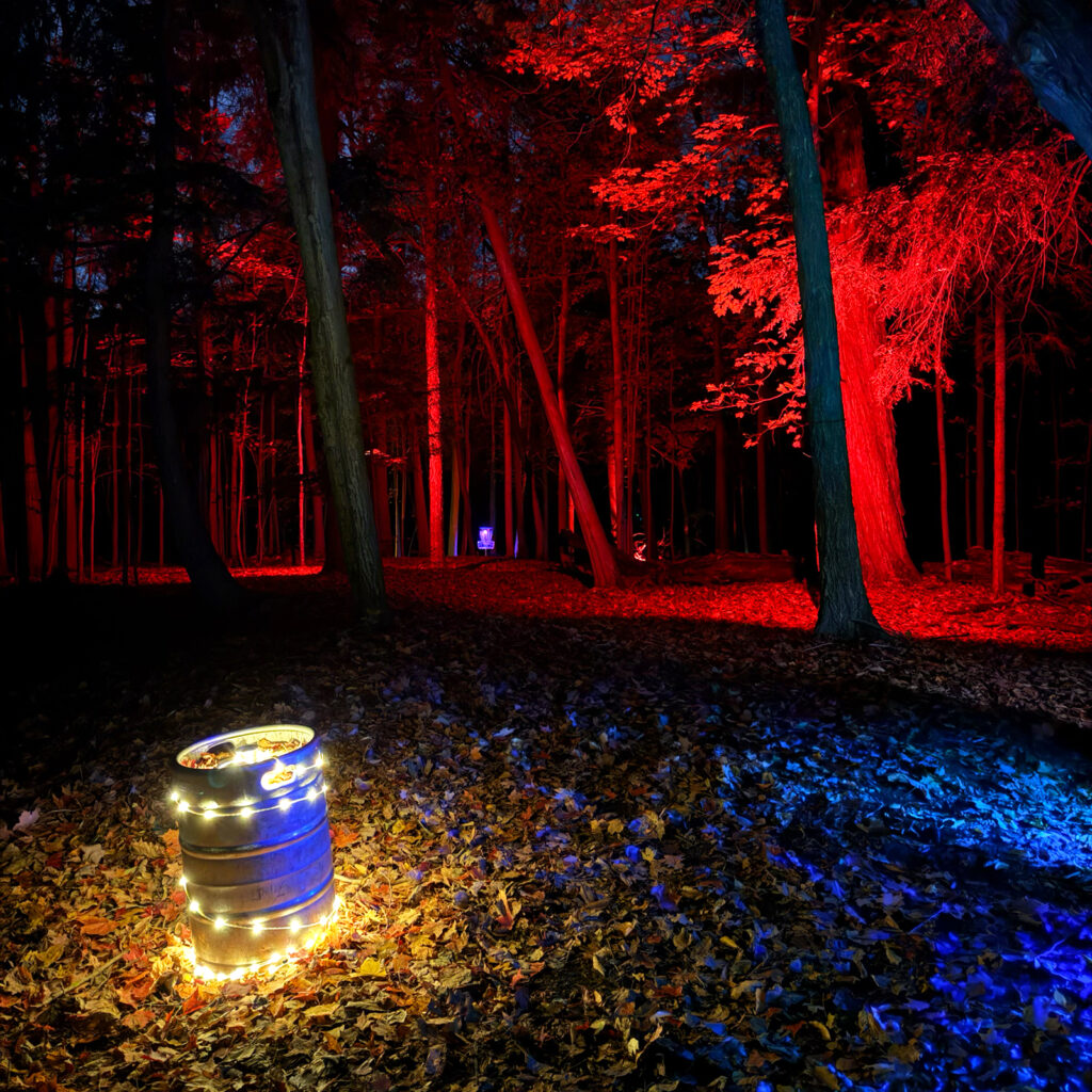 A string-light-wrapped keg glowing warm gold in a forest lit entirely red, with a basket visible far down the path