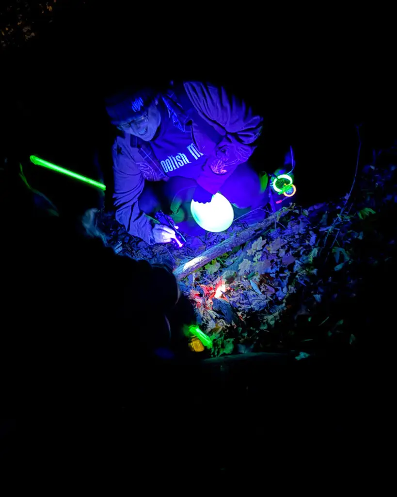 A player crouched down in the dark, lit in purple light, holding a glowing disc and surrounded by glow sticks