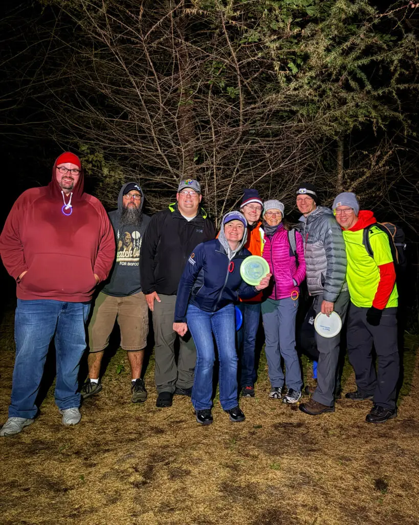 A large group of GLOWLOT players posing together in the dark woods, several holding glow discs