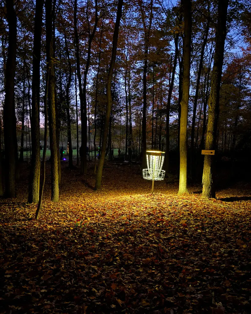 A warmly lit disc golf basket in the forest with an arrow sign pointing the way to the next hole