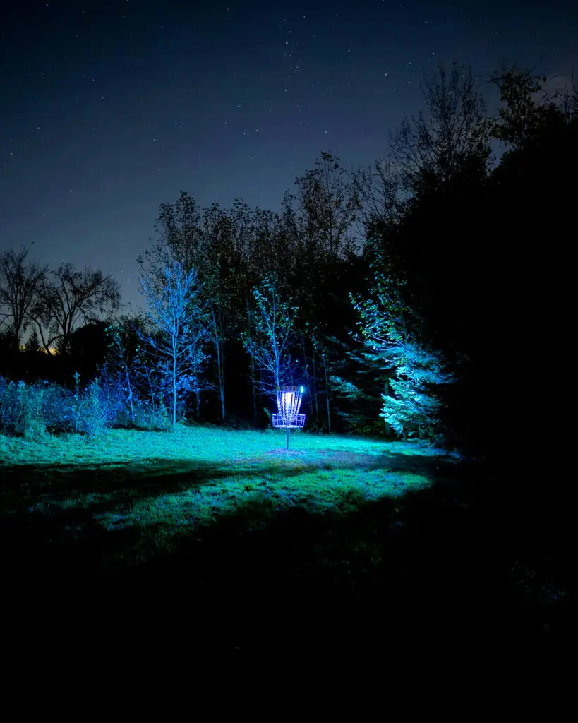 Hole 4's disc golf basket glowing blue-white in a grassy clearing surrounded by teal-lit trees