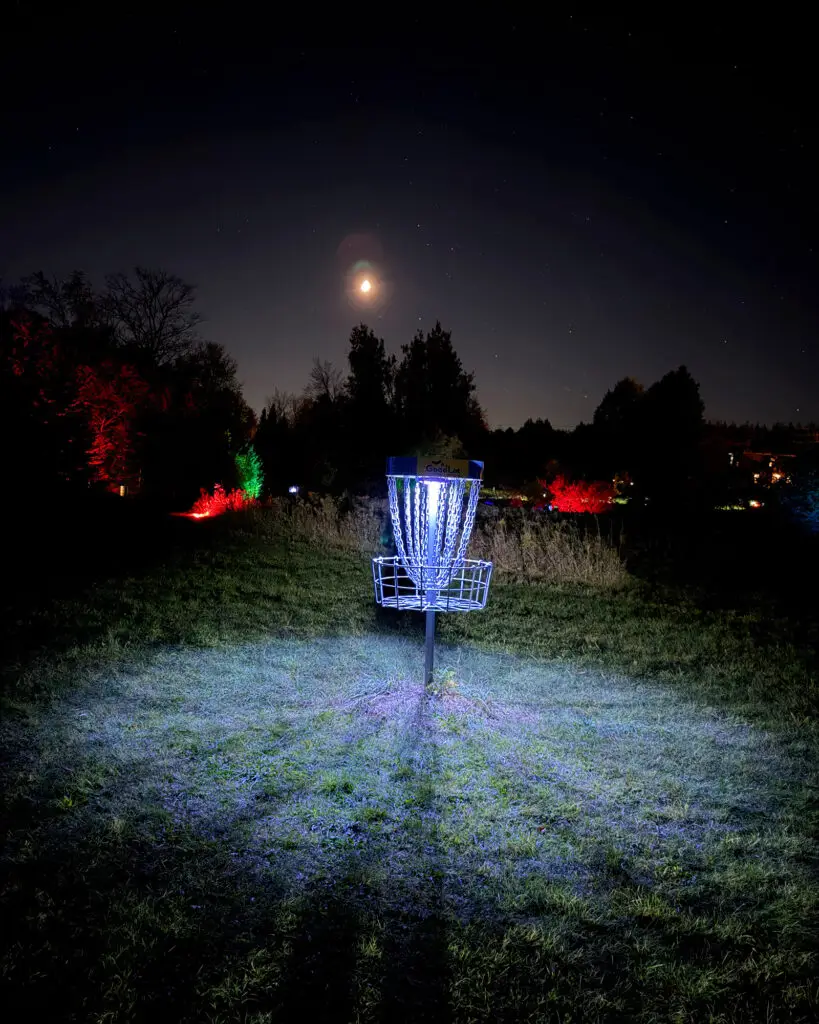A GoodLot disc golf basket glowing under a bright moon, with red and green lit trees in the background