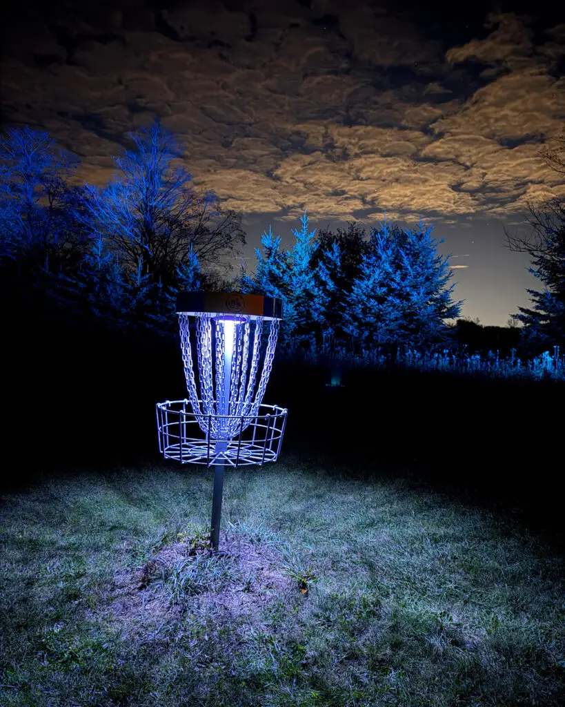 A glowing disc golf basket beneath the moon, with a horizon of red, green, and blue lit trees