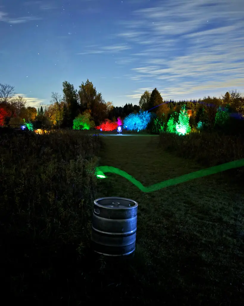 A glowing keg beside a lit green pathway leading toward a rainbow of illuminated trees at GLOWLOT