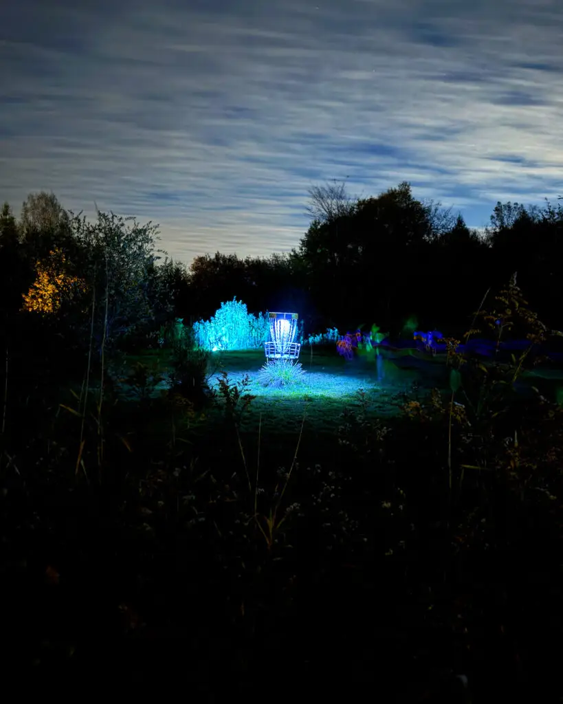 A teal-lit disc golf basket with players captured as glowing blurs moving past in the dark