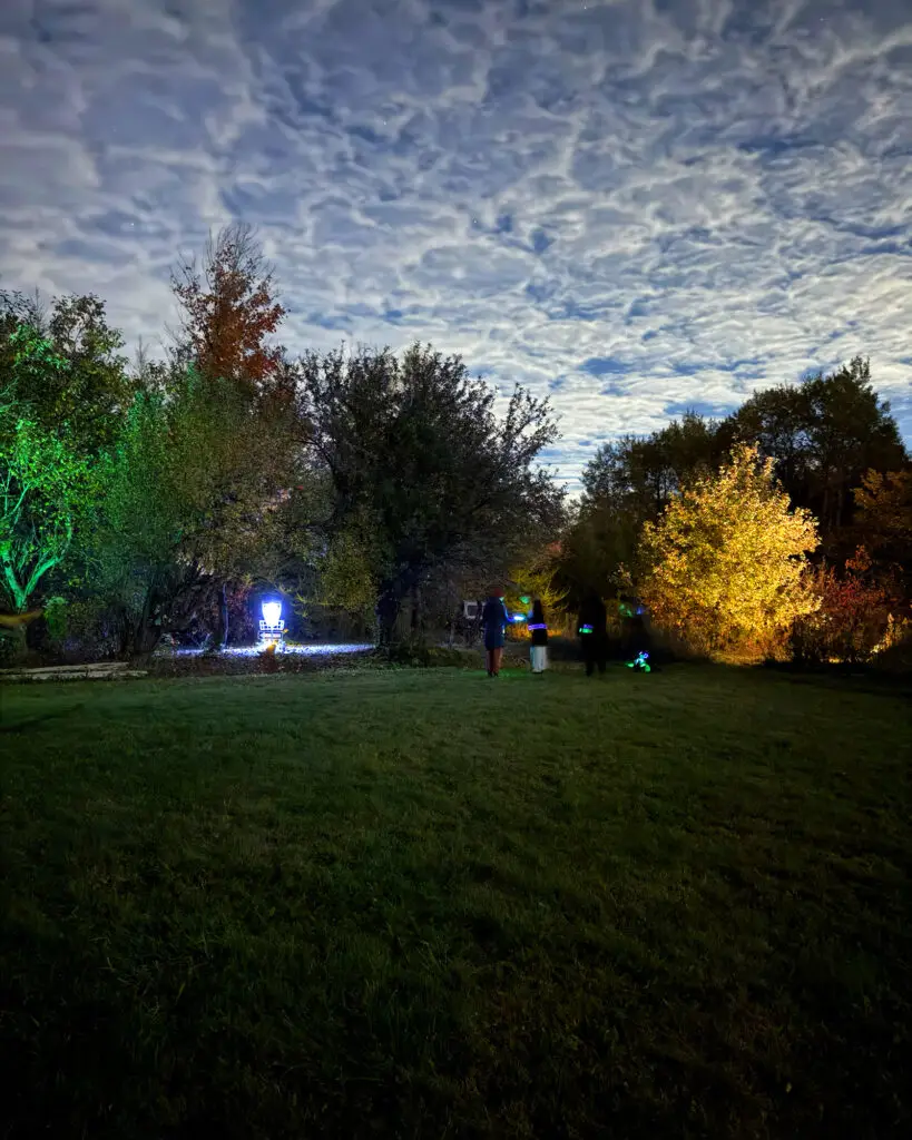 Three players standing in a grassy field at GoodLot with a glowing basket and yellow and green lit trees nearby