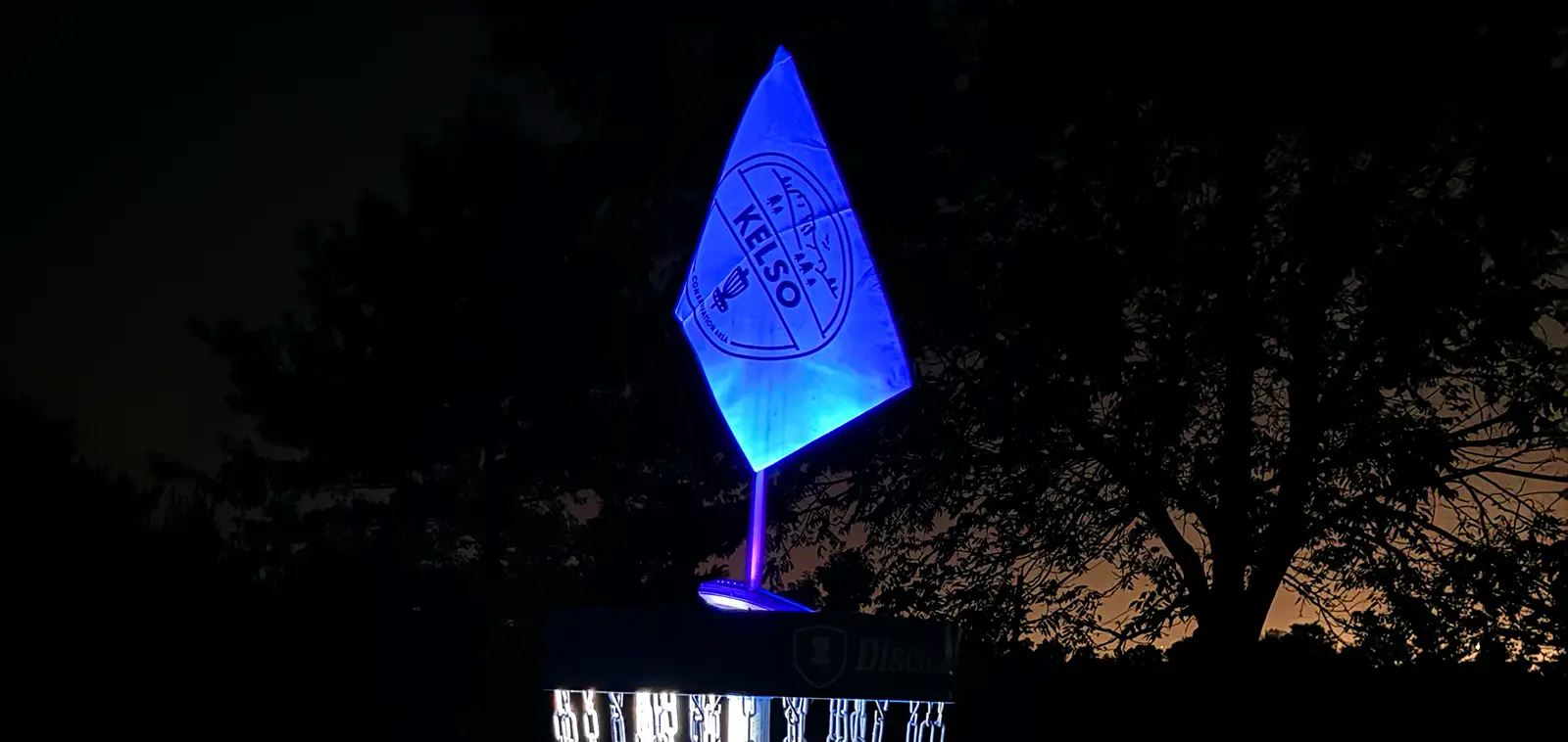 Close-up of a lit disc golf basket at night with a glowing blue flag showing the Kelso Conservation Area logo.