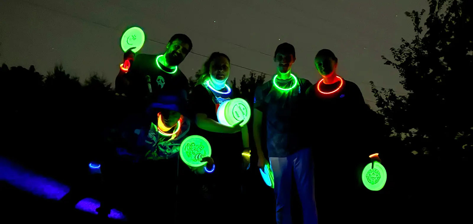 Five players pose at night wearing glow necklaces and holding glowing green discs under a starry sky.